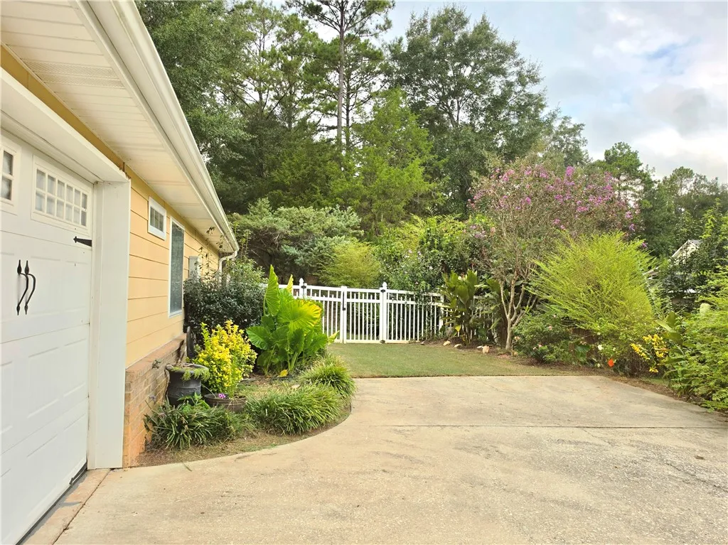 A spacious concrete driveway leads toward a fenced backyard surrounded by lush mature trees.