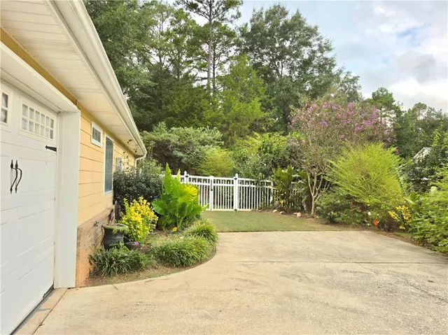 A spacious concrete driveway leads toward a fenced backyard surrounded by lush mature trees.