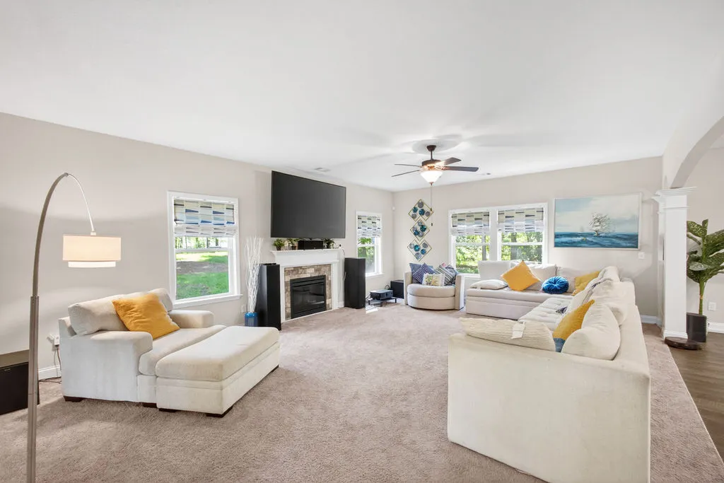 A well-lit gathering space features plush carpeting, a cozy fireplace, and several windows framing verdant outdoor views.