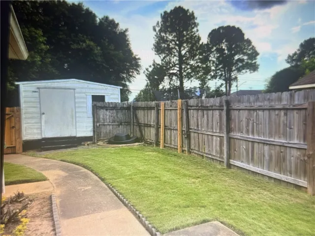A concrete walkway leads past a grassy lawn toward a wooden fence and storage shed.