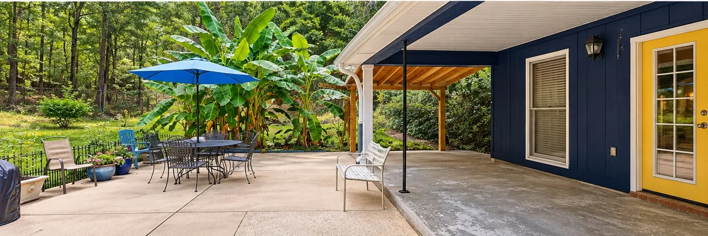 An expansive concrete patio connects seamlessly to a covered porch alongside lush, wooded backyard views.