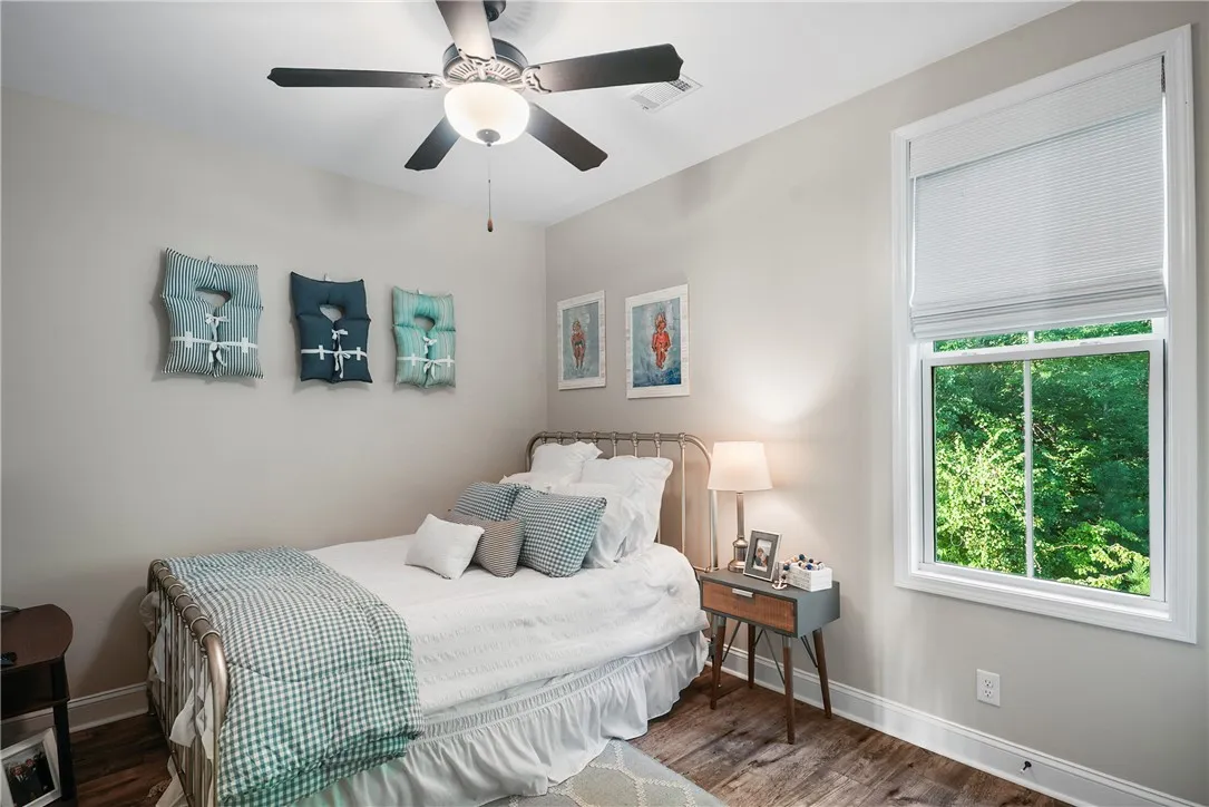 A bright bedroom showcases rich wood floors, high ceilings, and a tall window framing green views.