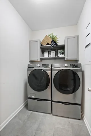 A functional utility space featuring modern side-by-side laundry units under matching cabinetry and tiled flooring.