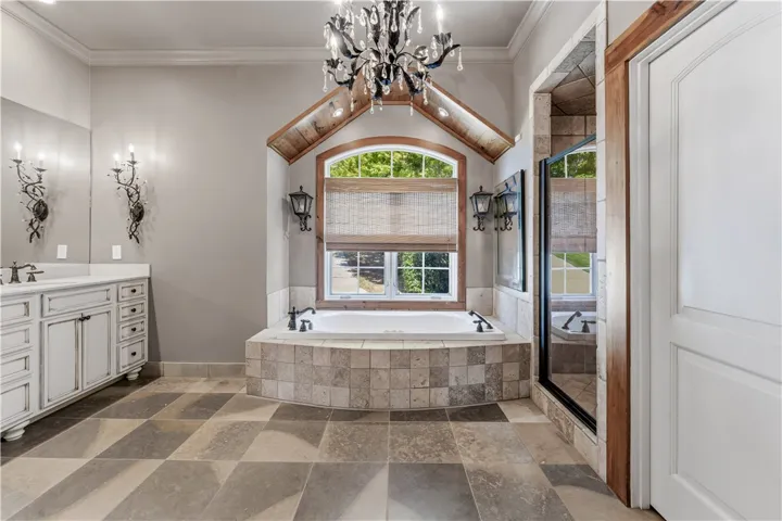 Ornate crystal lighting hangs above a tiled soaking tub set beneath a wood-accented arched window.