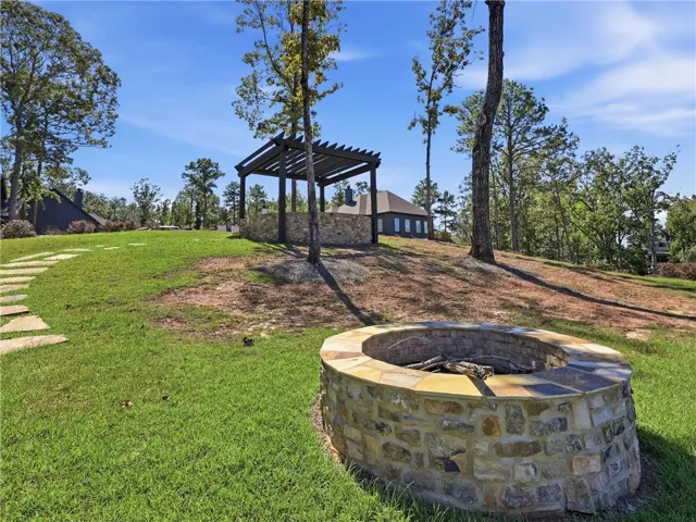 An inviting stone fire pit sits on a grassy slope beneath a shaded pergola structure.