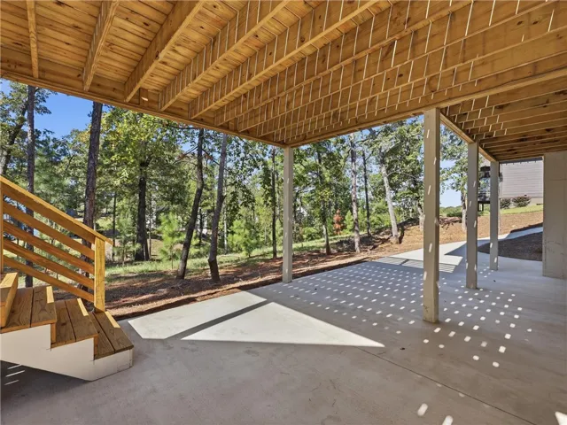 Shaded concrete patio features wooden stairs and overhead decking overlooking a wooded landscape.