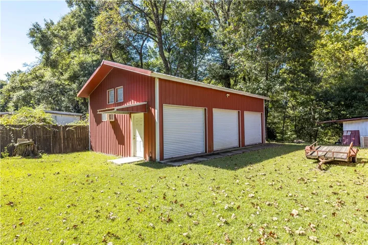 A spacious detached metal workshop stands on a grassy lawn bordered by mature trees.
