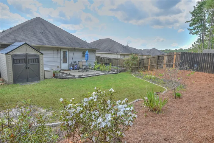 An enclosed backyard features a green lawn, garden beds, and a stone patio area.