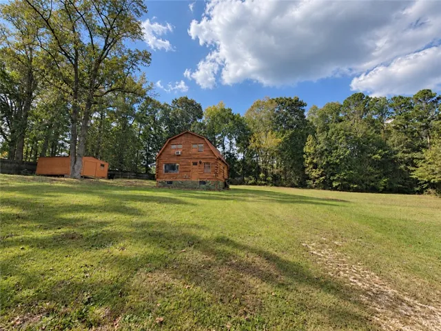 A charming log structure sits on a rolling green lawn bordered by mature trees.