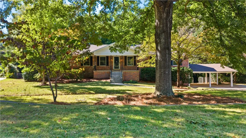 Mature trees shade a classic brick ranch home featuring an adjacent detached carport and manicured lawn.