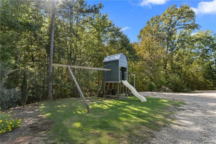 A wooden playset sits on a grassy lawn bordered by mature trees and a gravel drive.