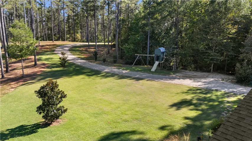 Mature trees frame a sprawling lawn and gravel driveway bordering a shaded play area.