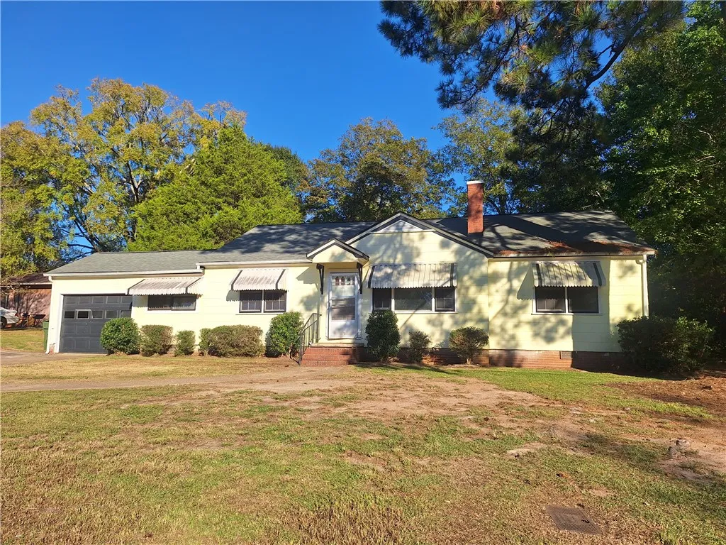 Mature trees frame a classic single-story home featuring a brick chimney and awnings over the windows.