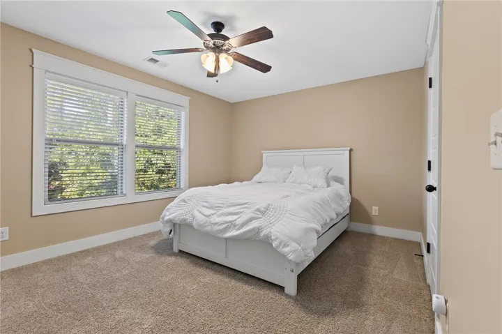 A spacious bedroom features plush carpeting and double windows framing leafy outdoor views.