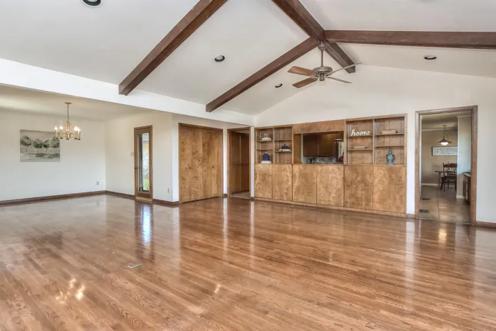 Unfurnished living room with beamed ceiling, high vaulted ceiling, ceiling fan with notable chandelier, wood finished floors, and baseboards