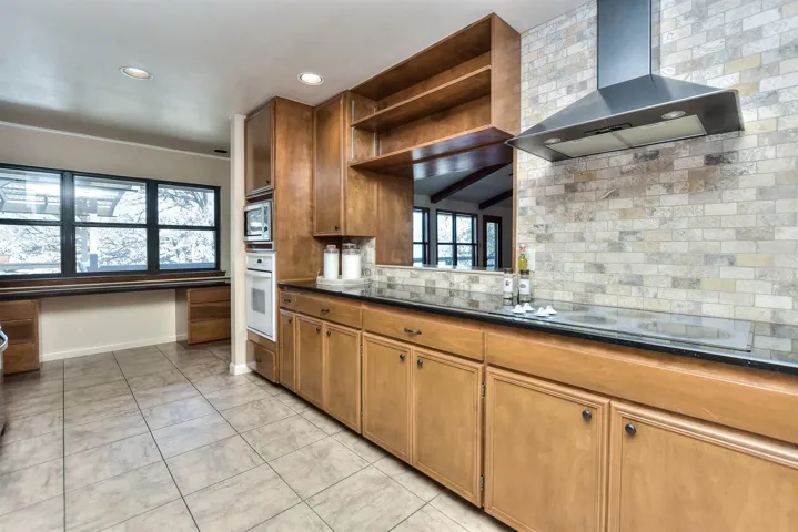 Kitchen with oven, stainless steel microwave, backsplash, wall chimney exhaust hood, and brown cabinetry