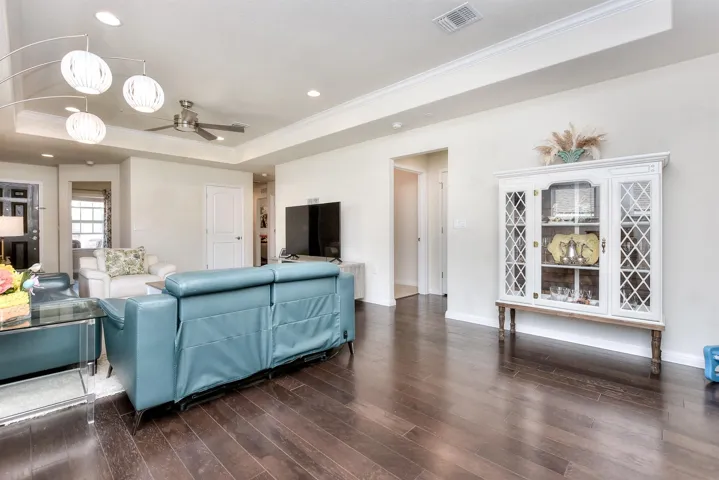 Living room with dark wood-style floors, visible vents, ceiling fan, and a tray ceiling