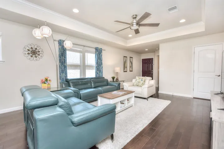 Living room with a tray ceiling, dark wood-style floors, visible vents, and ceiling fan