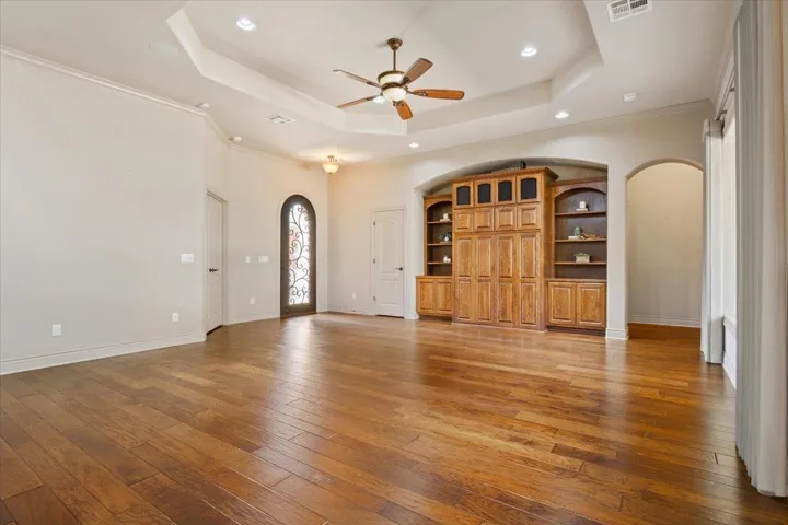 Unfurnished living room with arched walkways, light wood finished floors, a ceiling fan, a tray ceiling, and ornamental molding