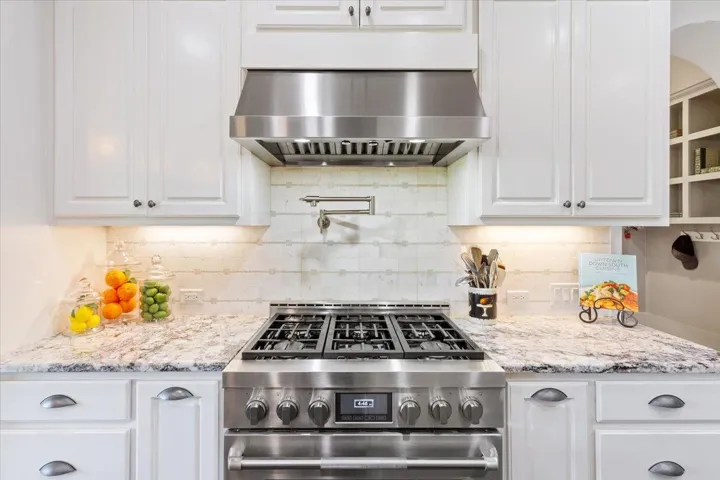 Kitchen featuring stainless steel, 6 burner range, exhaust hood, white cabinetry, and tasteful  natural stone backsplash...