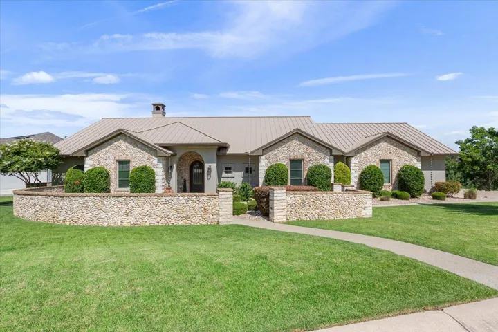 Single story home featuring a standing seam roof, a luscious front lawn, metal standing seam roof, and a half wall comprised of the same stone on the front of the home.