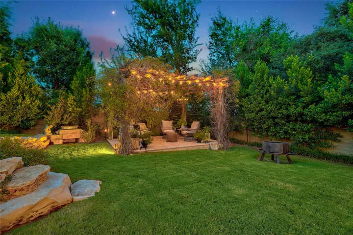 Lush green lawns and mature trees frame a stone-paved patio beneath a rustic wooden pergola.