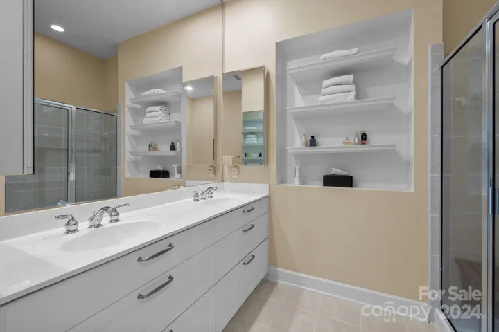 En-suite primary bathroom features expansive double-sink vanity with drawer storage, wall-mounted, mirrored medicine cabinets, tile flooring, built-in shelving.
