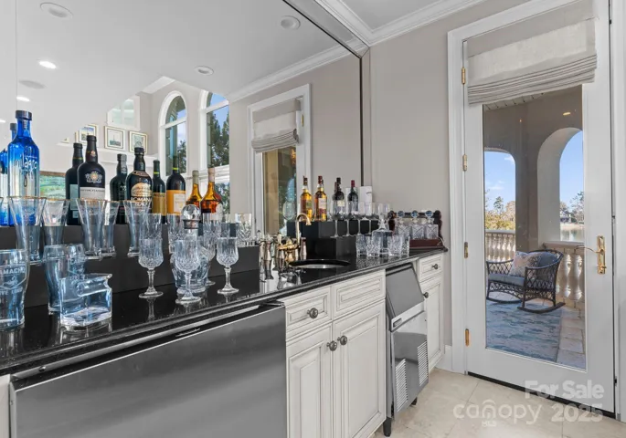 The bar area off the great room features a beverage refrigerator, ice maker, sink, and sleek black stone countertops. A door opens to the exterior covered patio, seamlessly connecting indoor and outdoor living spaces.