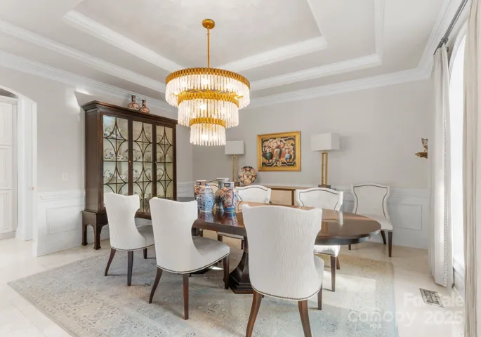 The elegant dining room boasts tray ceilings, a stunning chandelier, and a large window that frames the view of the front of the home, creating a sophisticated and welcoming atmosphere.