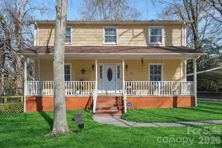 Front exterior daytime view of this charming two-story home featuring a welcoming covered porch, classic white railing, and brick foundation detail. Fresh green lawn enhances curb appeal, complemented by mature trees and a curved walkway leading to the front entry. Spacious driveway with carport offers convenient covered parking. A warm, inviting elevation that highlights the home’s character, outdoor living space, and well-maintained presentation.
