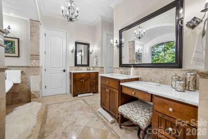 The thoughtfully curated primary bathroom includes heated floors, limestone with inset tiles, separate Carrera marble sinks plus a vanity area, custom framed mirrors, beautiful lighting and a Sunrise Specialty dual slipper tub with a large window view of the private side yard.