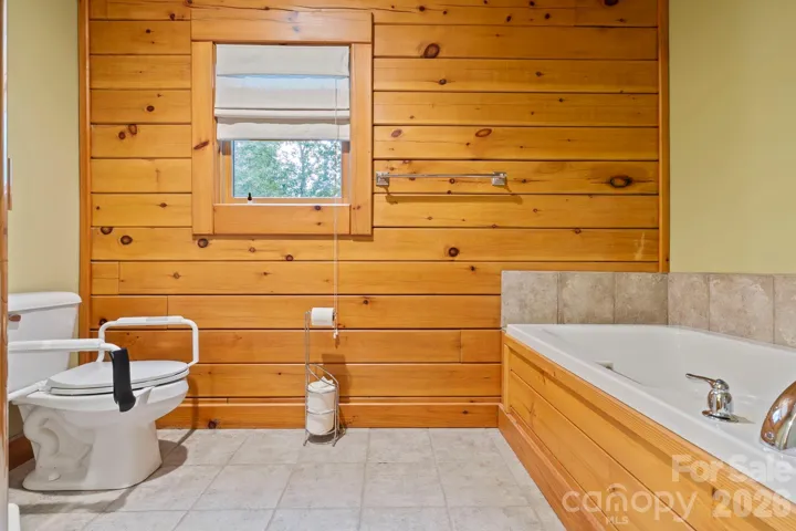 Full bath with soaking tub, wood-paneled walls, and window bringing in natural light and serene wooded views.