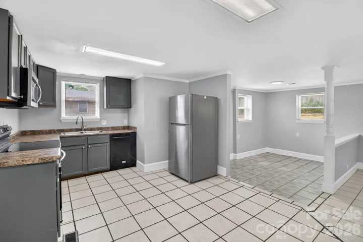 This spacious and bright kitchen offers a clean, modern aesthetic with a functional layout perfect for home chefs and entertainers alike.Kitchen HighlightsModern Cabinetry: Sophisticated charcoal-grey cabinetry provides ample storage and a sleek contrast against the light grey walls.Updated Appliances: Equipped with a stainless steel refrigerator, microwave, and a convenient black dishwasher.Clean Finishes: Features durable laminate countertops with a natural stone look and a deep double-basin stainless steel sink situated under a bright window.Easy Maintenance: Large-format tiled flooring runs throughout the kitchen, offering a durable surface that is easy to clean.Space & LayoutThe open-concept design flows seamlessly into a versatile sun-drenched dining area or breakfast nook. This additional space features secondary tiling and large windows, creating a bright, airy atmosphere for morning coffee or casual family meals.This versatile space is move-in ready and waiting for your pe
