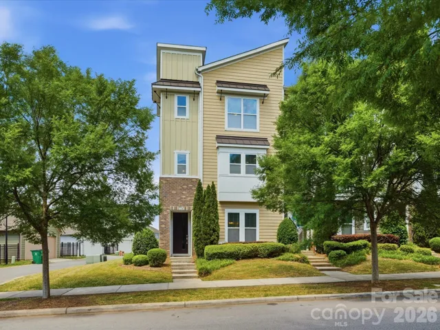 Striking three-story townhome with cream board-and-batten siding, brick-accented entry, and lush mature shade trees delivering instant curb appeal in a sought-after walkable Brightwalk community.