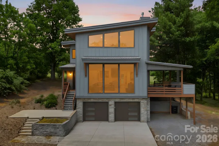 Striking modern mountain home with dramatic floor-to-ceiling windows, clean architectural lines, and warm interior lighting that creates a stunning evening presence. The covered side deck and wooded setting add to the home’s private retreat feel.