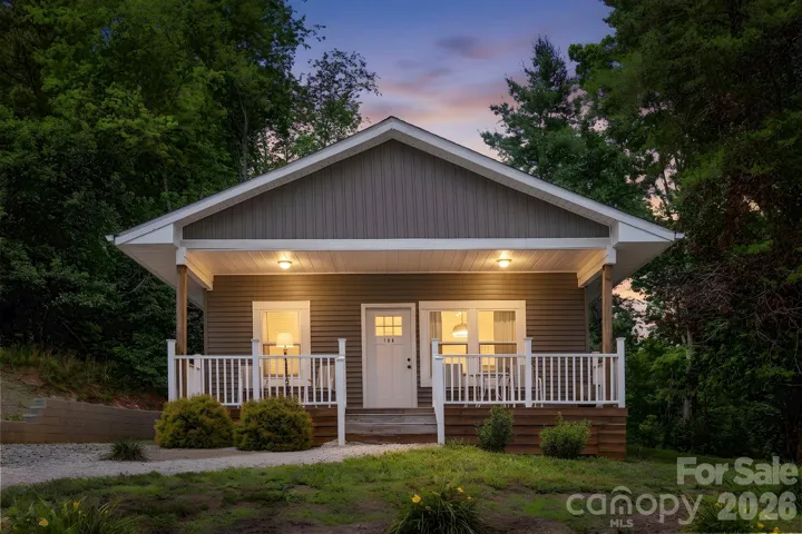 Charming cottage-style exterior showcased at dusk, with warm interior lighting, a covered front porch, and a peaceful wooded setting.