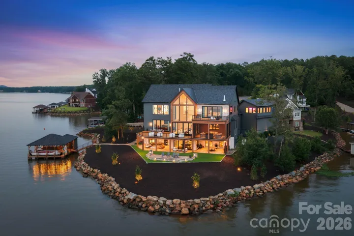 Aerial view showcasing the lake-facing side of the property, including the boathouse, turfed grounds, lower-level patio, decks, and walls of glass