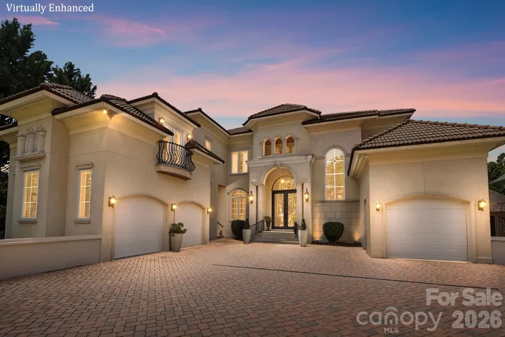 An impressive twilight view showcases the home’s grand Mediterranean-inspired façade, highlighted by warm architectural lighting and a dramatic arched entry. Multiple rooflines, elegant stucco detailing, wrought-iron balcony accents, expansive windows, and a spacious motor court create a striking sense of arrival, while three garage bays provide exceptional convenience.
