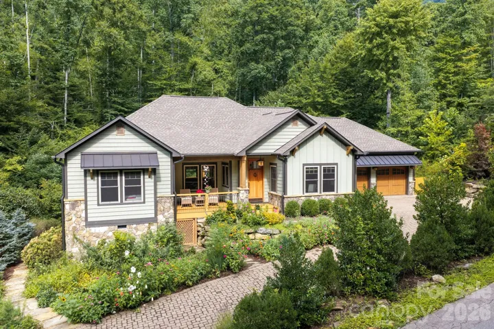 Beautifully crafted mountain home nestled against a lush wooded backdrop, featuring board-and-batten and shake siding, natural stone accents, metal awnings, and a welcoming covered front porch. Paver walkway winds through vibrant, professionally landscaped perennial gardens to the front entry and attached two-car garage.