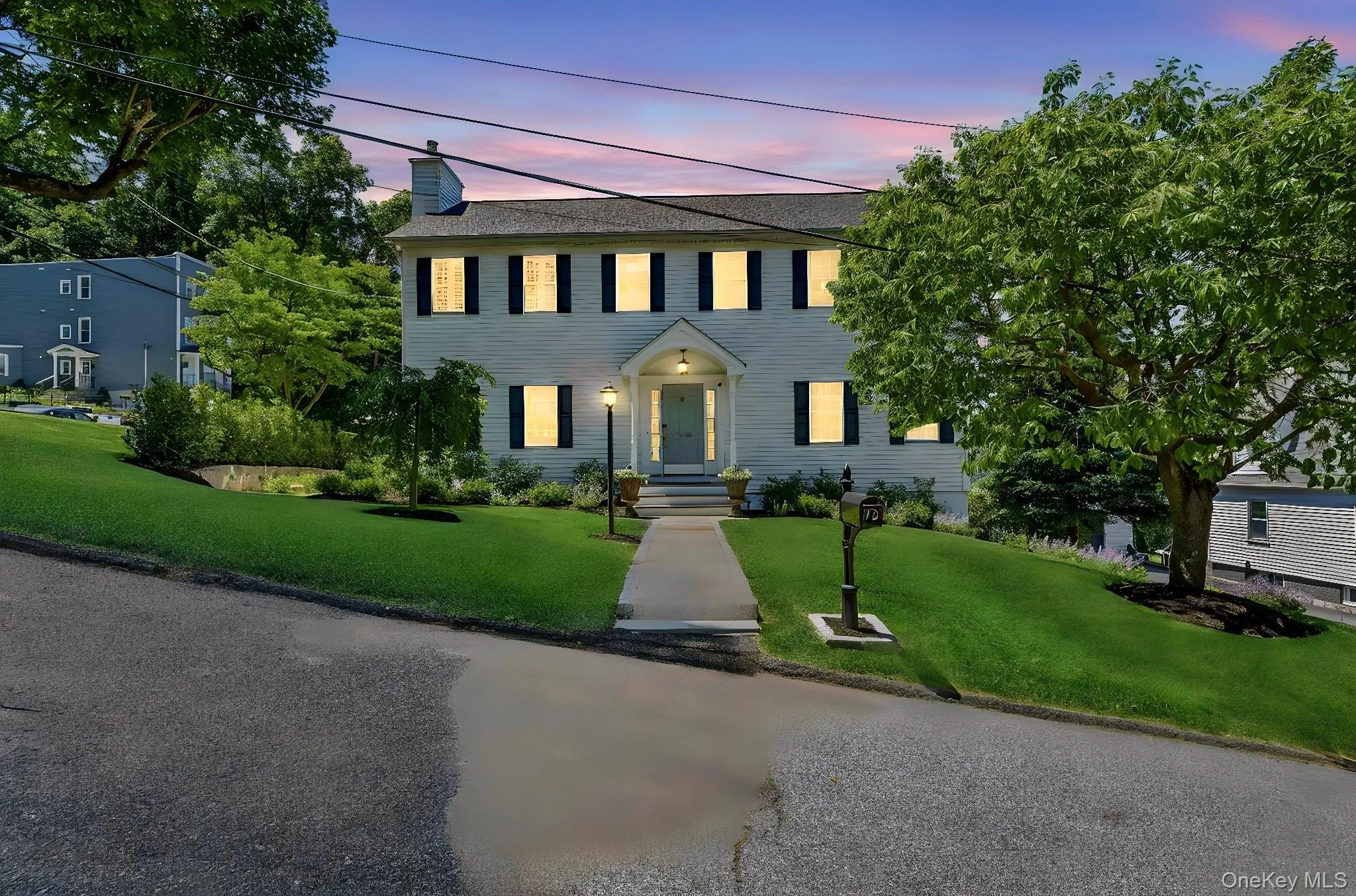 Traditional two-story residence featuring a covered portico entry, black window shutters, and a sloped front lawn with mature landscaping Traditional two-story residence featuring a covered portico entry, black window shutters, and a sloped front lawn with mature landscaping