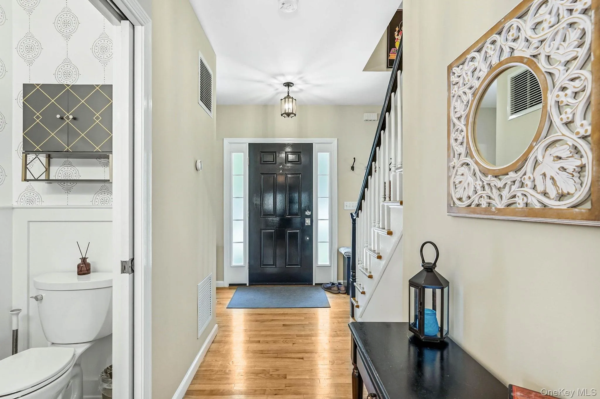 Bright entryway featuring wood-finish flooring, a black paneled front door with sidelights, and a staircase with white spindles and a dark handrail Bright entryway featuring wood-finish flooring, a black paneled front door with sidelights, and a staircase with white spindles and a dark handrail