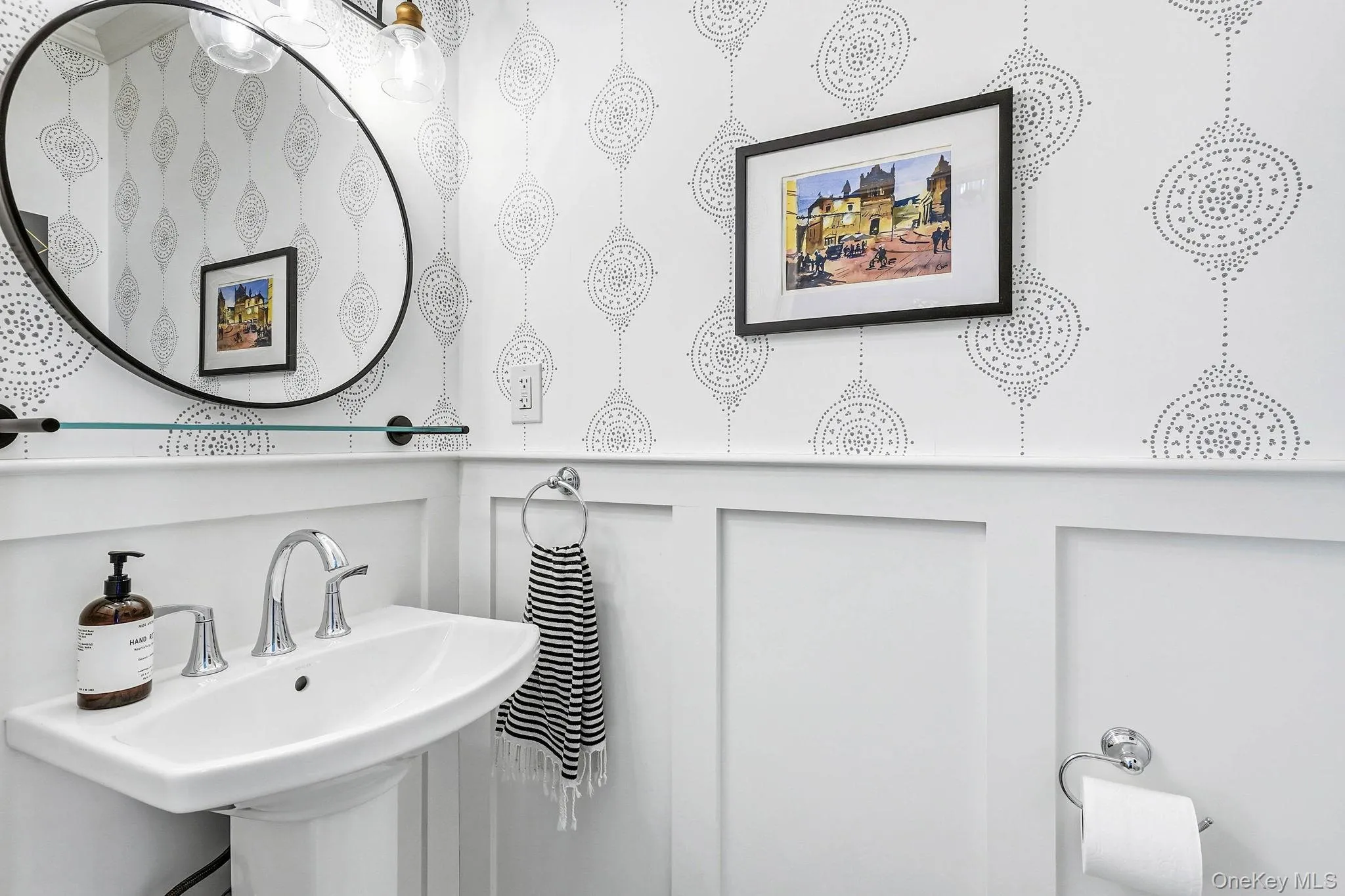 Pedestal sink featuring a chrome faucet, black-framed oval mirror, board and batten wainscoting, and patterned wallpaper Pedestal sink featuring a chrome faucet, black-framed oval mirror, board and batten wainscoting, and patterned wallpaper