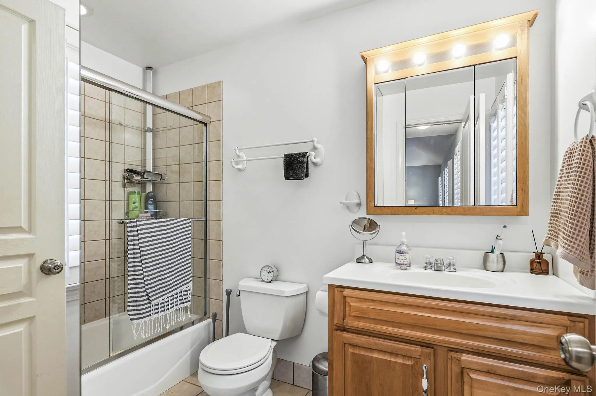 Full bathroom featuring a shower-tub combination with sliding glass doors, neutral tile surround, and a wood vanity with integrated sink Full bathroom featuring a shower-tub combination with sliding glass doors, neutral tile surround, and a wood vanity with integrated sink