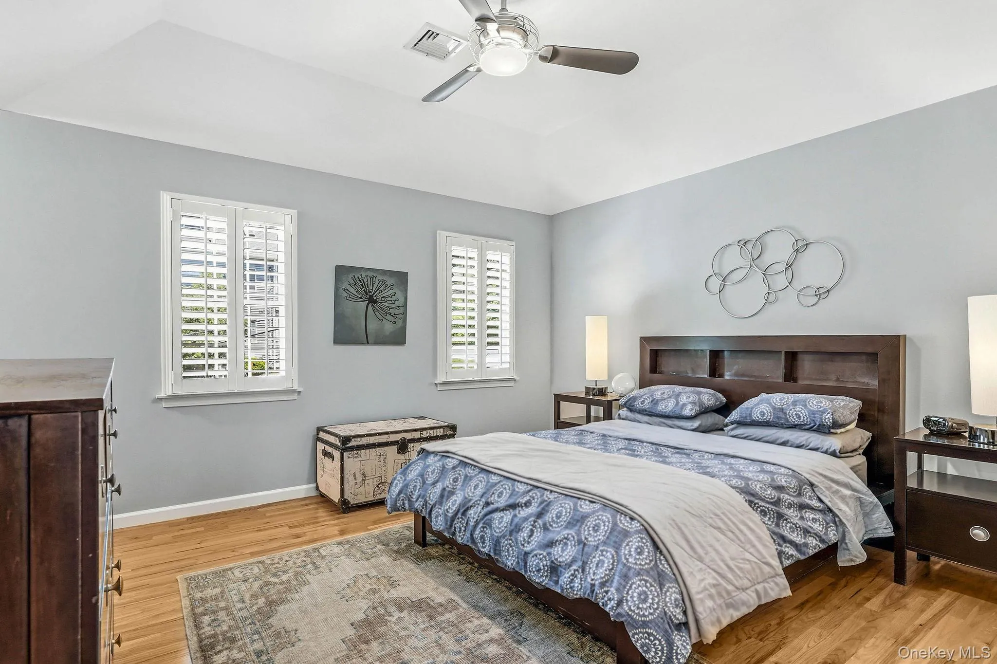 Room featuring wood-finish flooring, two windows with plantation shutters, a vaulted ceiling, a ceiling fan with integrated lighting, and a neutral wall color Room featuring wood-finish flooring, two windows with plantation shutters, a vaulted ceiling, a ceiling fan with integrated lighting, and a neutral wall color