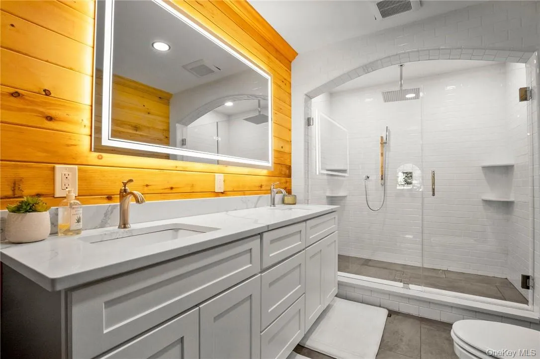 Bathroom with toilet, dual bowl vanity, wood walls, tile patterned flooring, and a shower with door Bathroom with toilet, dual bowl vanity, wood walls, tile patterned flooring, and a shower with door