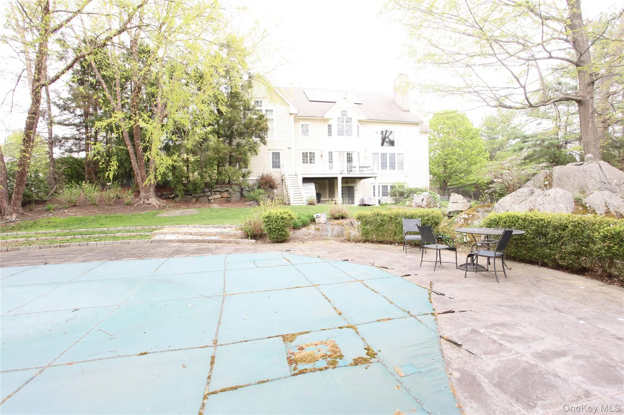 Spacious rear yard featuring a covered inground pool, concrete patio, natural rock formations, mature trees, and a multi-level home with a rear deck Spacious rear yard featuring a covered inground pool, concrete patio, natural rock formations, mature trees, and a multi-level home with a rear deck