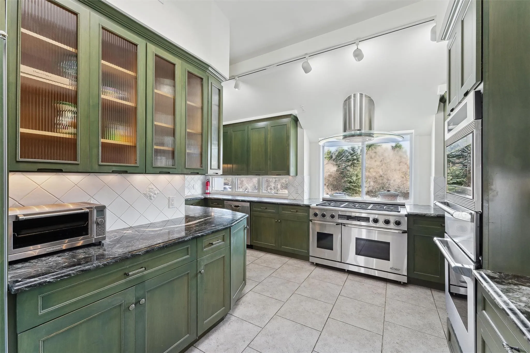 Kitchen with light tile patterned flooring, dark stone counters, green cabinets, island range hood, and stainless steel appliances Kitchen with light tile patterned flooring, dark stone counters, green cabinets, island range hood, and stainless steel appliances