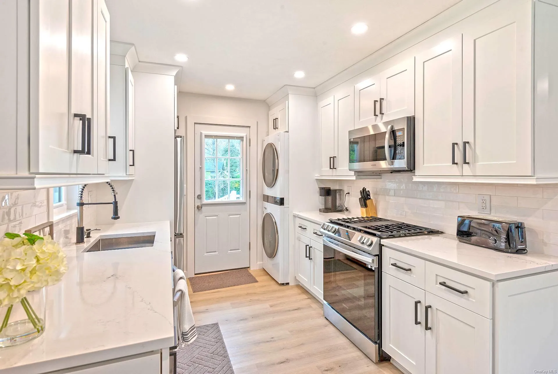 Kitchen featuring stainless steel appliances, stacked washer / drying machine, white cabinetry, decorative backsplash, and light wood finished floors Kitchen featuring stainless steel appliances, stacked washer / drying machine, white cabinetry, decorative backsplash, and light wood finished floors
