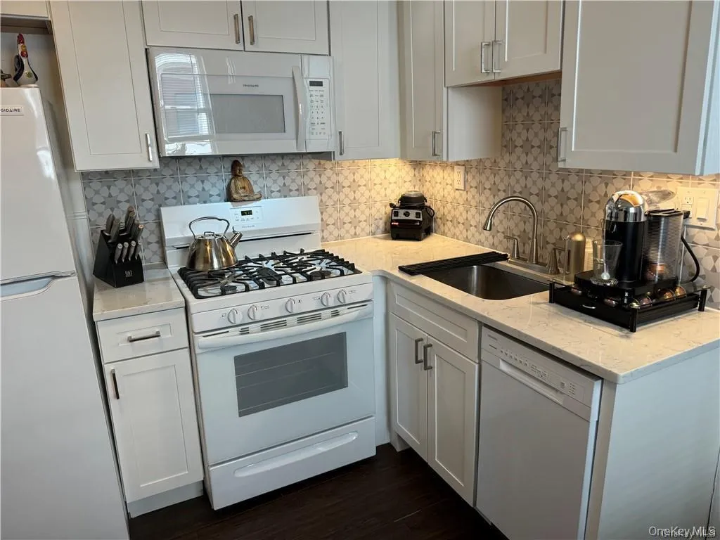 Kitchen with white appliances, decorative backsplash, dark wood finished floors, light stone counters, and white cabinets Kitchen with white appliances, decorative backsplash, dark wood finished floors, light stone counters, and white cabinets