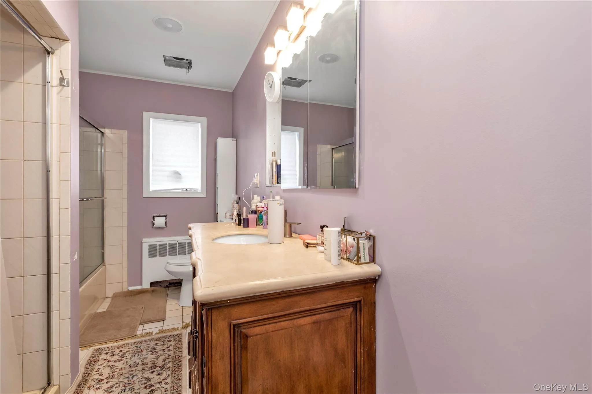 Full bathroom with vanity, shower / bath combination with glass door, tile patterned floors, toilet, and radiator heating unit Full bathroom with vanity, shower / bath combination with glass door, tile patterned floors, toilet, and radiator heating unit